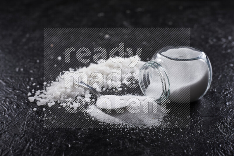 A glass jar full of table salt with some sea salt crystals beside it on a black background