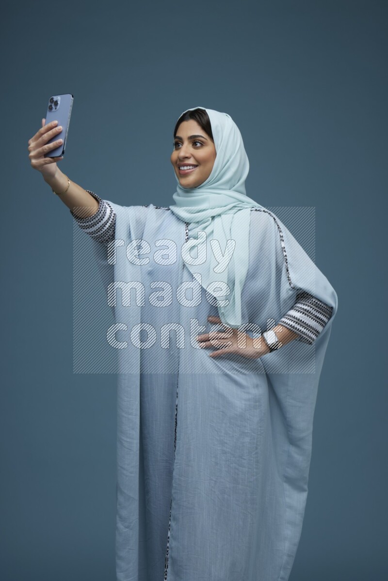A Saudi woman Taking a Selfie on a blue background wearing a blue Abaya with hijab