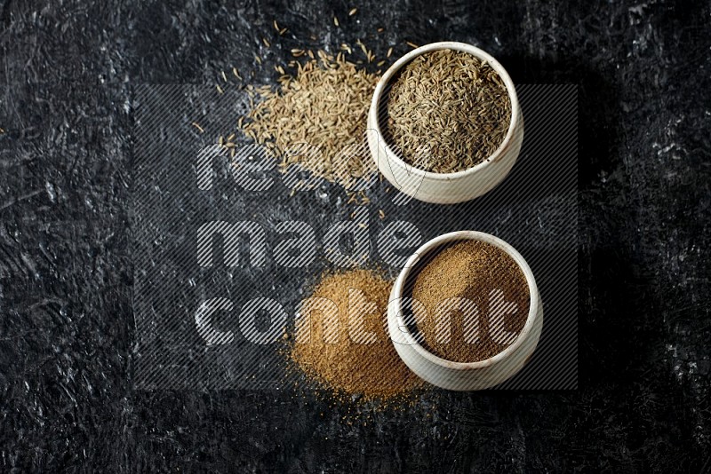 2 beige bowls, one full of cumin powder and the other full of seeds on a textured black flooring