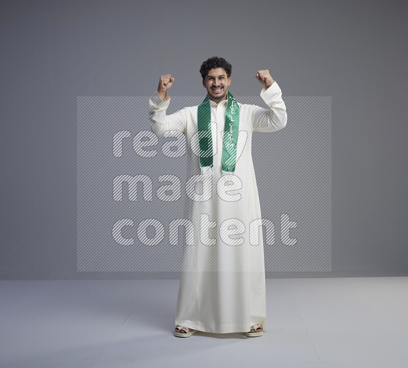 A Saudi man standing wearing thob and Saudi flag scarf on gray background