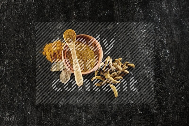 A wooden bowl and a wooden spoon full of turmeric powder with dried turmeric fingers on textured black flooring