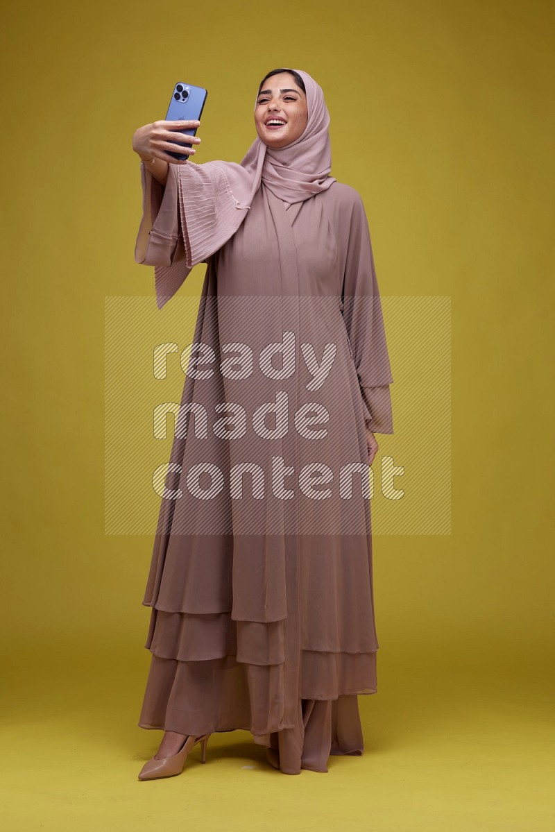 A woman taking a Selfie on a Yellow Background wearing Brown Abaya with Hijab