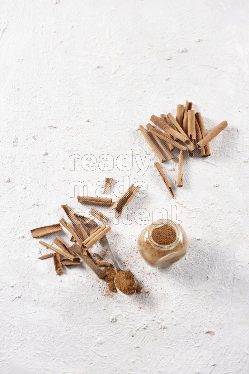 Herbal glass jar full cinnamon powder and a metal spoon surrounded by cinnamon sticks on a white background