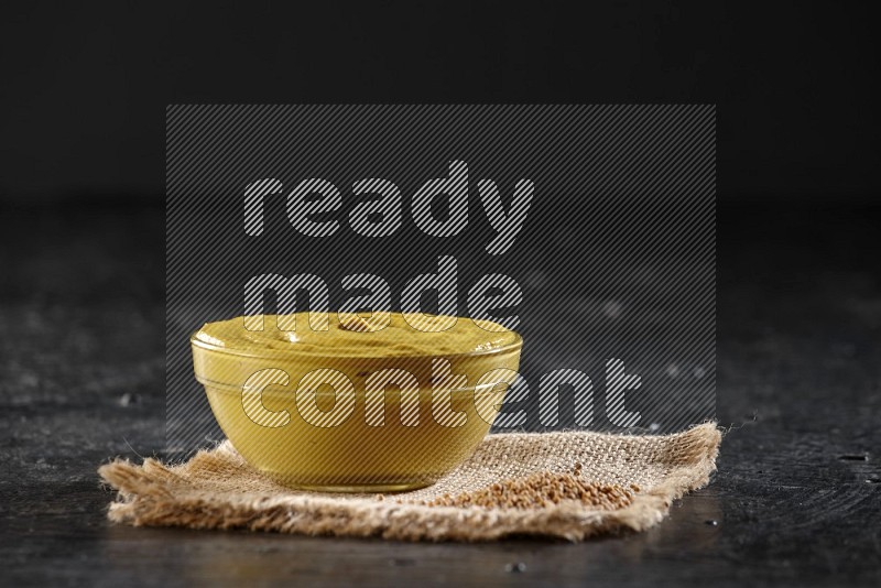 A glass bowl full of mustard paste set on a burlap piece with some mustard seeds on a textured black flooring in different angles