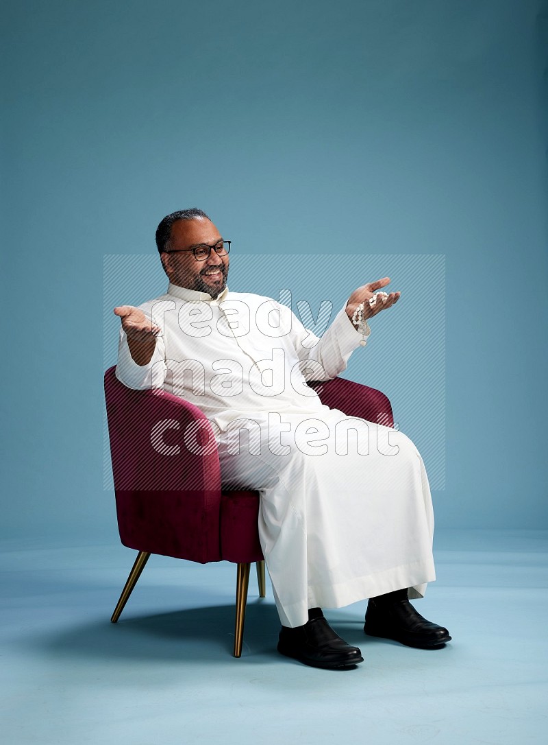 Saudi Man without shimag sitting on chair Interacting with the camera on blue background