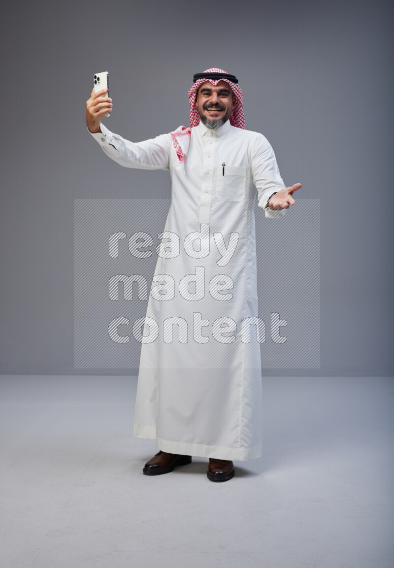 Saudi man Wearing Thob and red Shomag standing taking selfie on Gray background