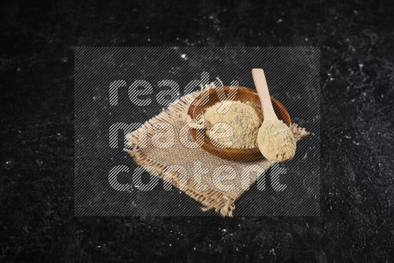 A wooden bowl full of ground ginger powder with a wooden spoon on it all on a burlap fabric on black background