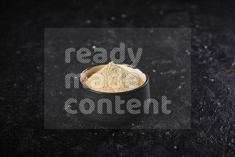 A black pottery bowl full of ground ginger powder on black background