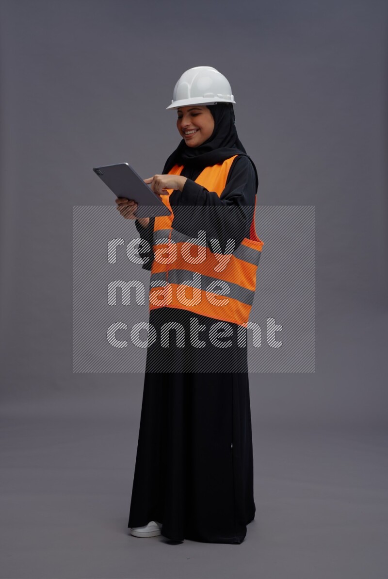 Saudi woman wearing Abaya with engineer vest standing working on tablet on gray background