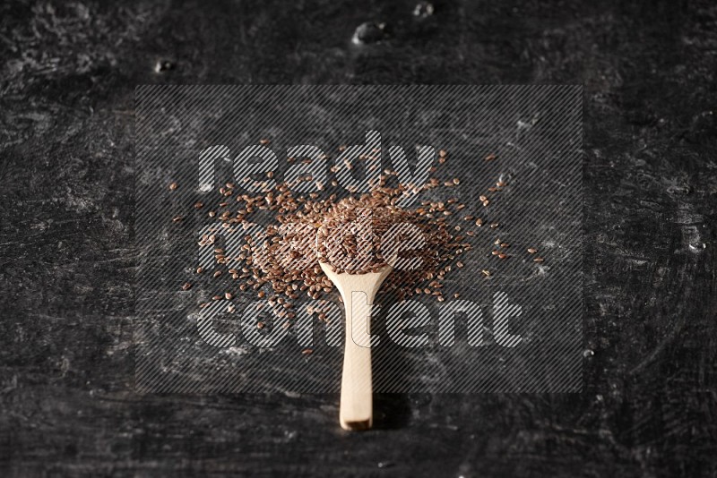 A wooden spoon full of flaxseeds and surrounded by seeds on a textured black flooring