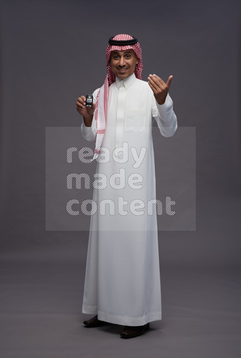 Saudi man wearing thob and shomag standing holding key car on gray background