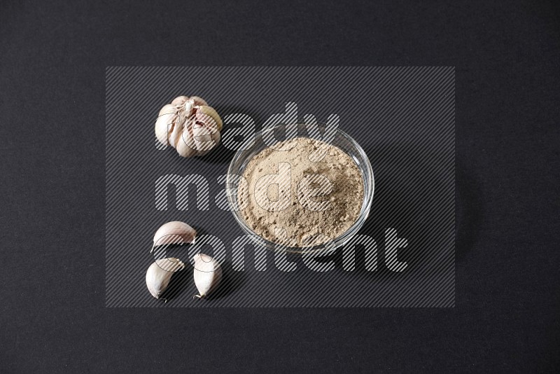 A glass bowl full of garlic powder with garlic bulb and some cloves beside it on a black flooring