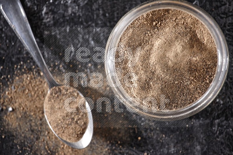 A glass jar full of black pepper powder and a metal spoon full of powder on a textured black flooring