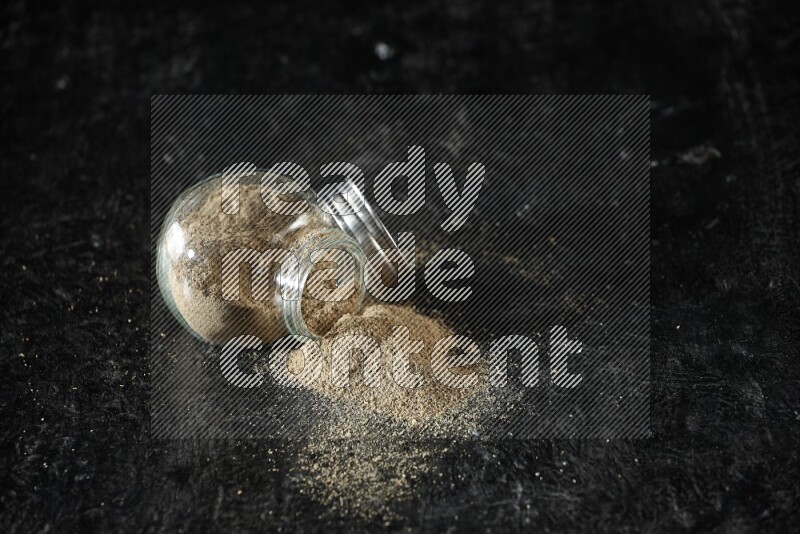 A flipped glass spice jar full of cardamom powder on textured black flooring