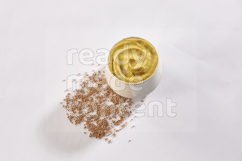 A beige pottery bowl full of mustard paste with mustard seeds underneath on white flooring