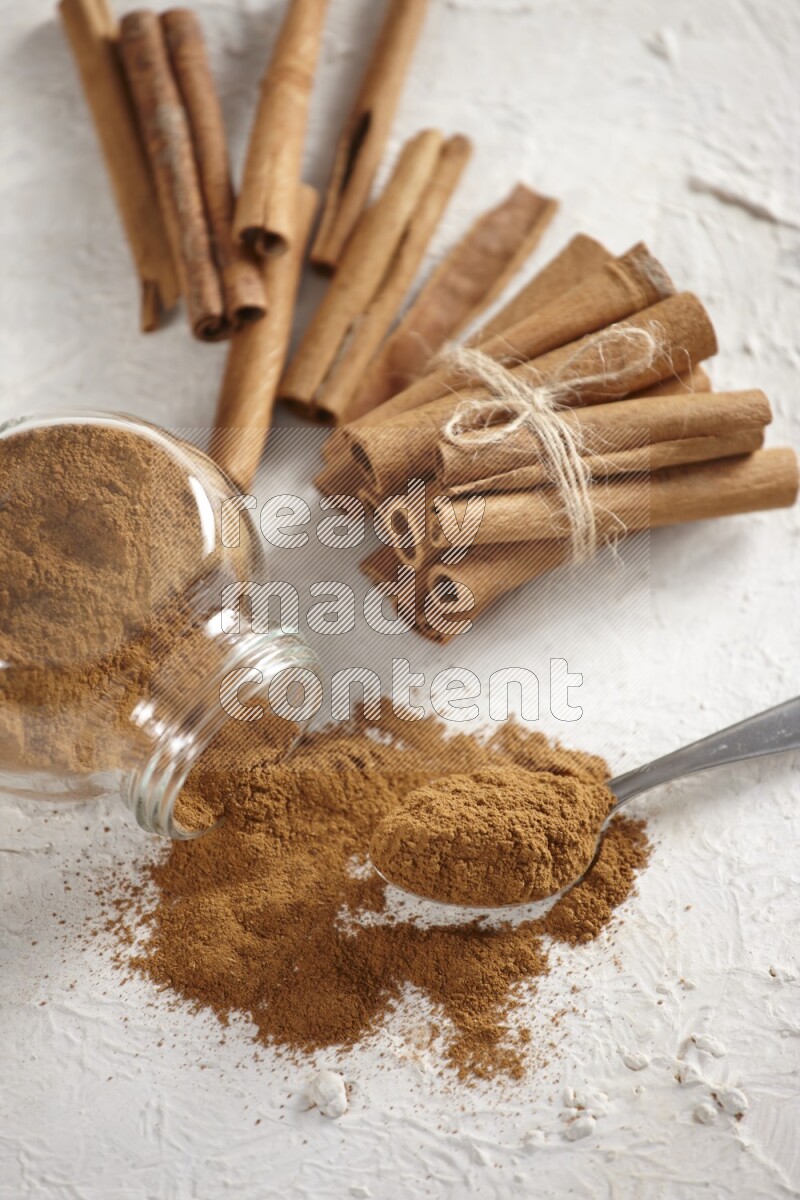Flipped herbs glass jar full of cinnamon powder with a metal spoon full of powder and cinnamon sticks on a textured white background