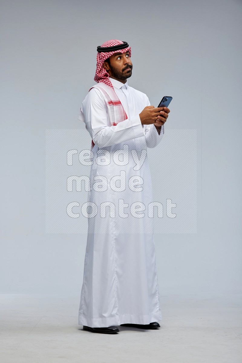 Saudi man Wearing Thob and shomag standing texting on phone on Gray background