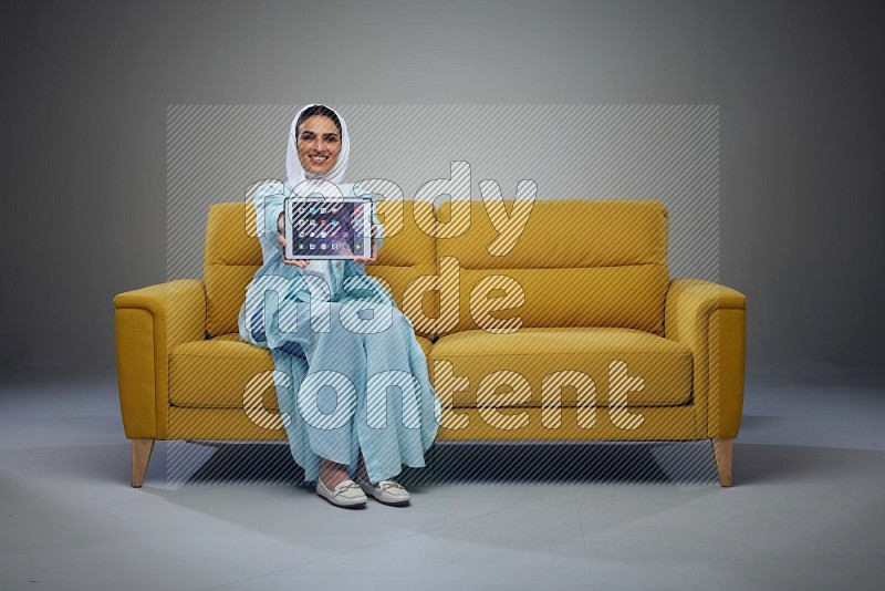 A Saudi woman wearing a light blue Abaya and white head scarf sitting on a yellow sofa and showing her tablet's screen while pointing to it eye level on a grey background