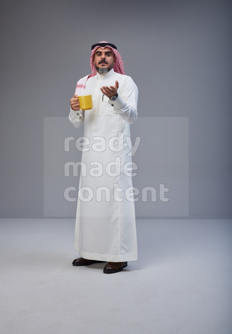Saudi man Wearing Thob and red Shomag standing holding mug on Gray background