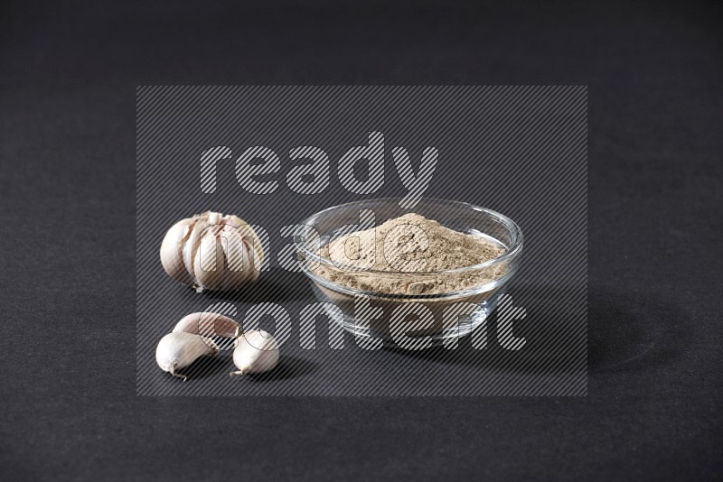 A glass bowl full of garlic powder with garlic bulb and some cloves beside it on a black flooring