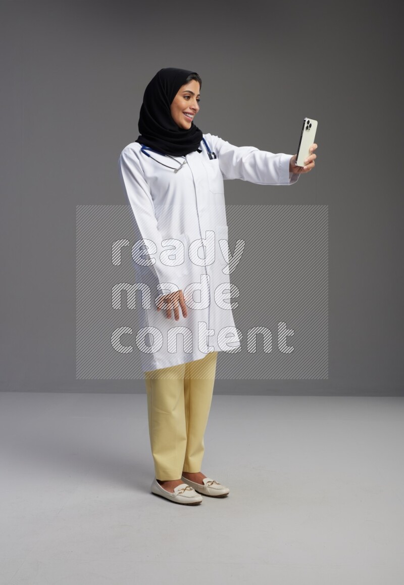 Saudi woman wearing lab coat with stethoscope standing taking selfie on Gray background