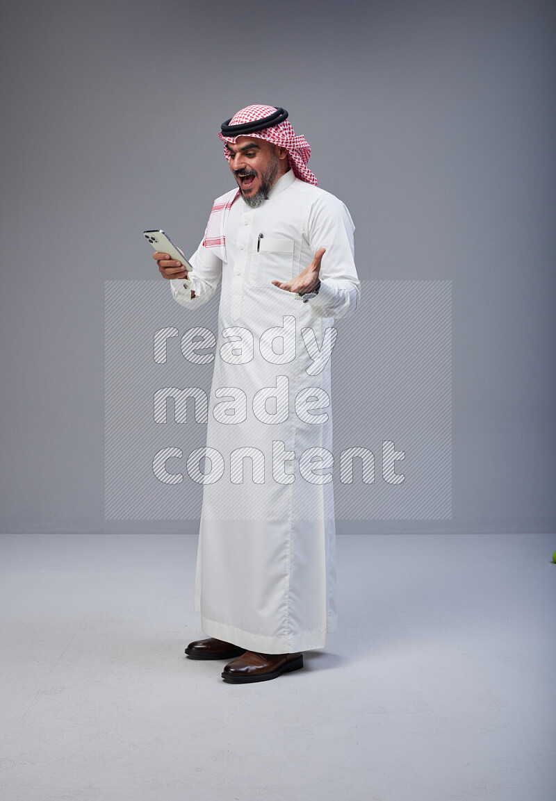 Saudi man Wearing Thob and red Shomag standing texting on phone on Gray background