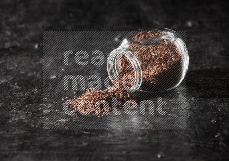 A glass spice jar full of flax flipped and seeds spreaded out on a textured black flooring in different angles
