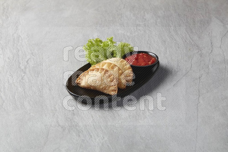 Three fried sambosas in an oval shaped black plate and a red sauce in a black round ramekin on a gray background