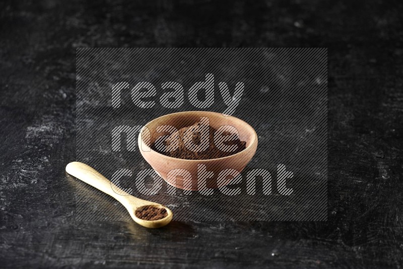 A wooden bowl and wooden spoon full of cloves powder on textured black flooring