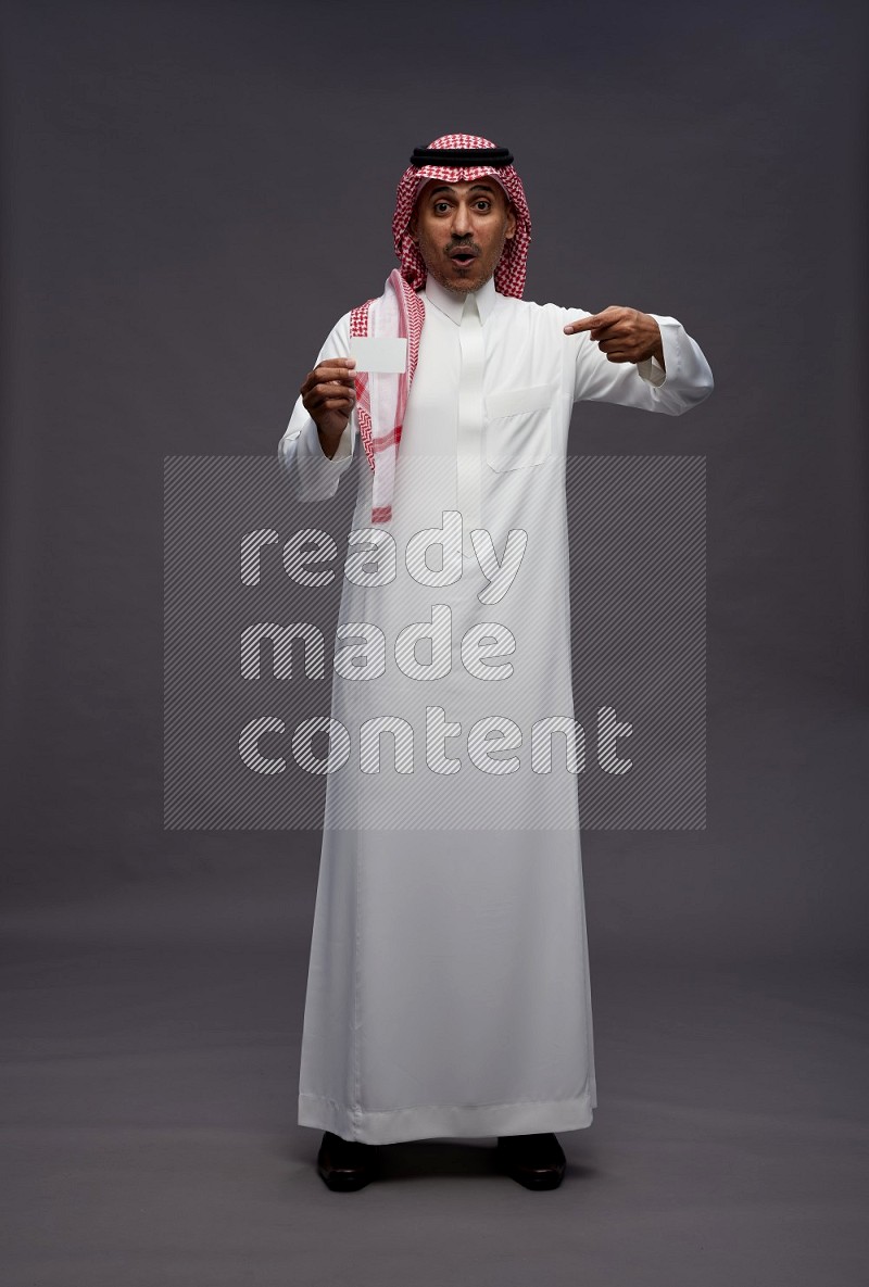 Saudi man wearing thob and shomag standing holding ATM card on gray background