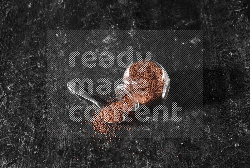 A glass spice jar full of garden cress seeds flipped and seeds spread out with a metal spoon full of the seeds on a textured black flooring