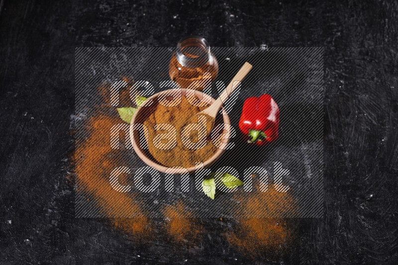 A wooden bowl full of ground paprika powder with a glass jar beside it and a red bell pepper on black background