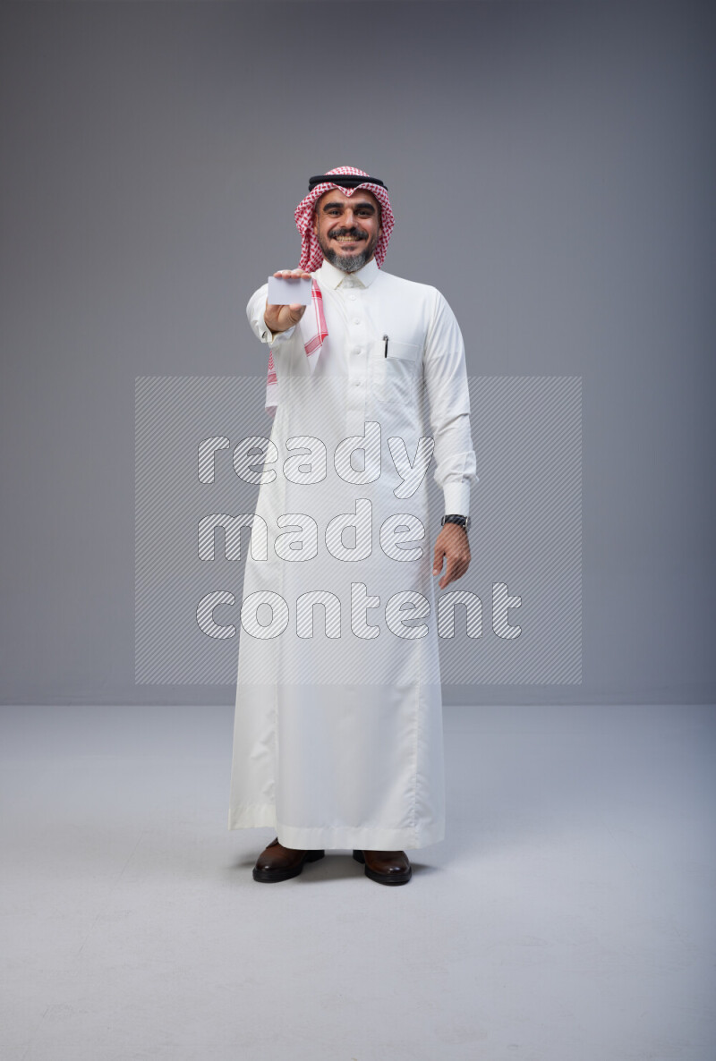 Saudi man Wearing Thob and red Shomag standing holding ATM card on Gray background