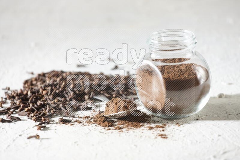 A glass spice jar and a metal spoon full of cloves powder and cloves spread on textured white flooring