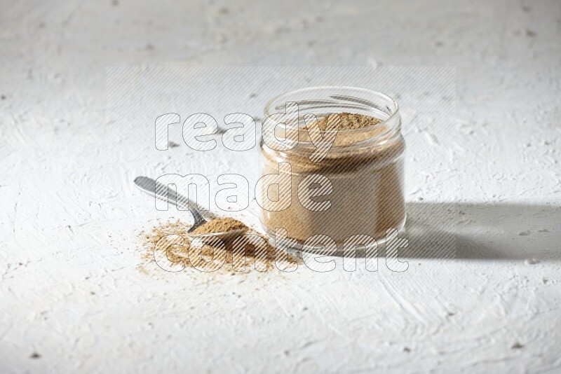A glass jar and a metal spoon full of cumin powder on textured white flooring