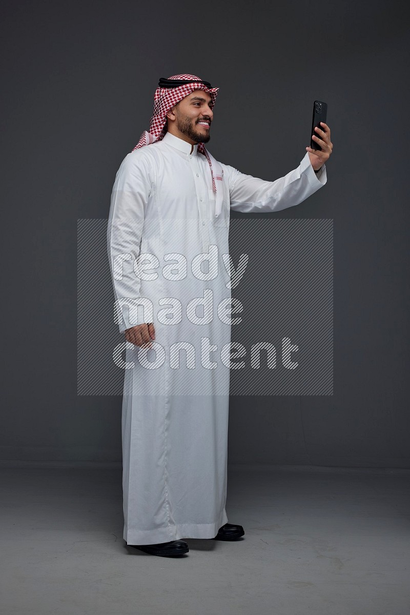 A Saudi man wearing Thobe and Shamgh taking selfie with his phone eye level on a gray background