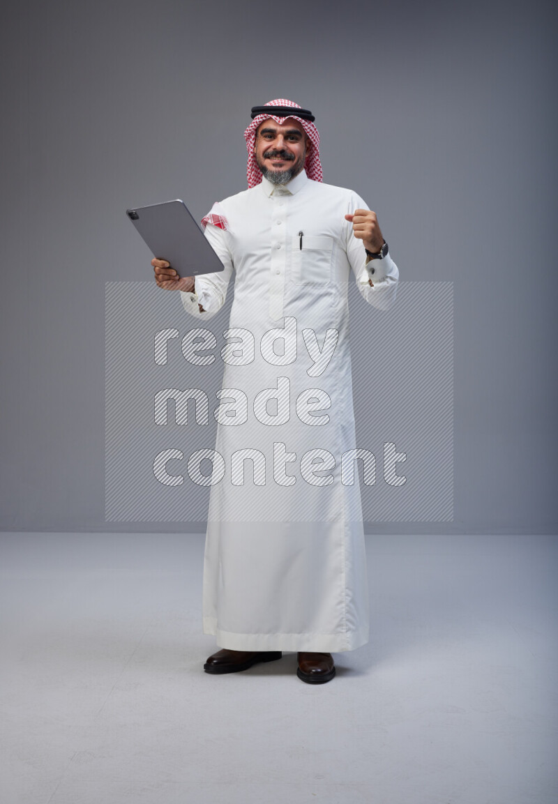 Saudi man Wearing Thob and red Shomag standing working on tablet on Gray background