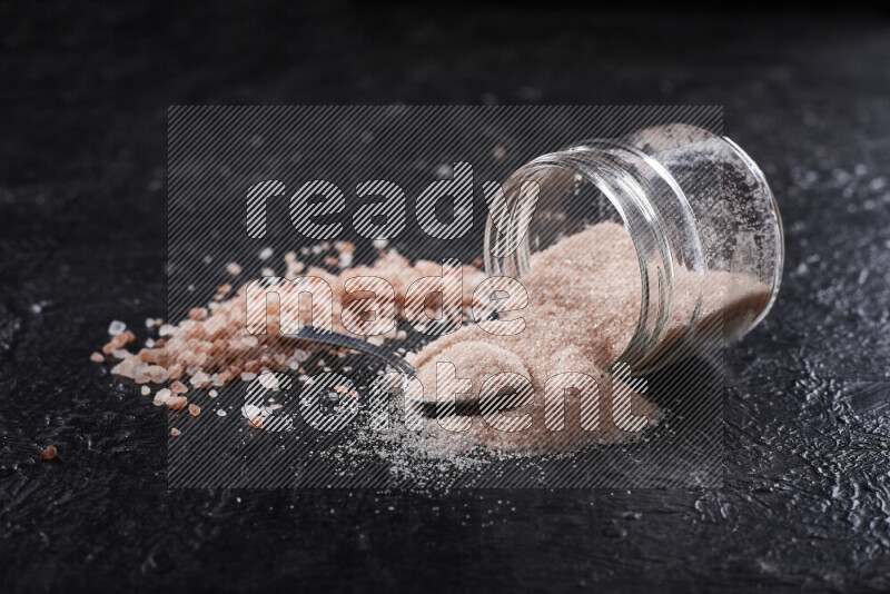 A glass jar full of fine himalayan salt with some himalayan crystals beside it on a black background