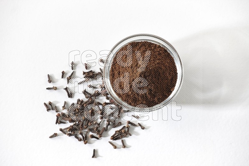 A glass bowl full of cloves powder and cloves grains spread on white flooring