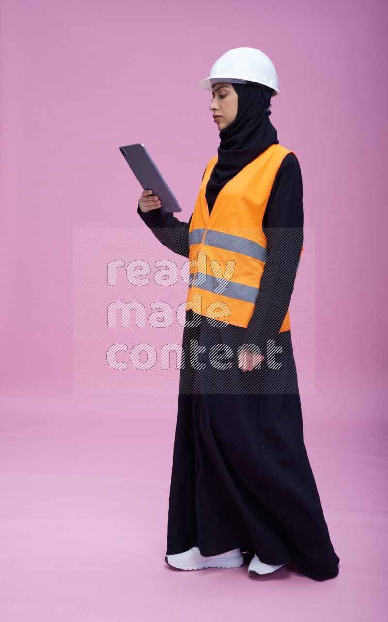 Saudi woman wearing Abaya with engineer vest and helmet standing working on tablet on pink background