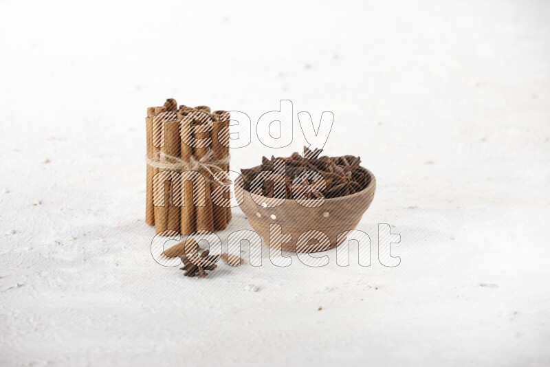 A stacked and bounded cinnamon sticks and a wooden bowl full of star anise on a white background