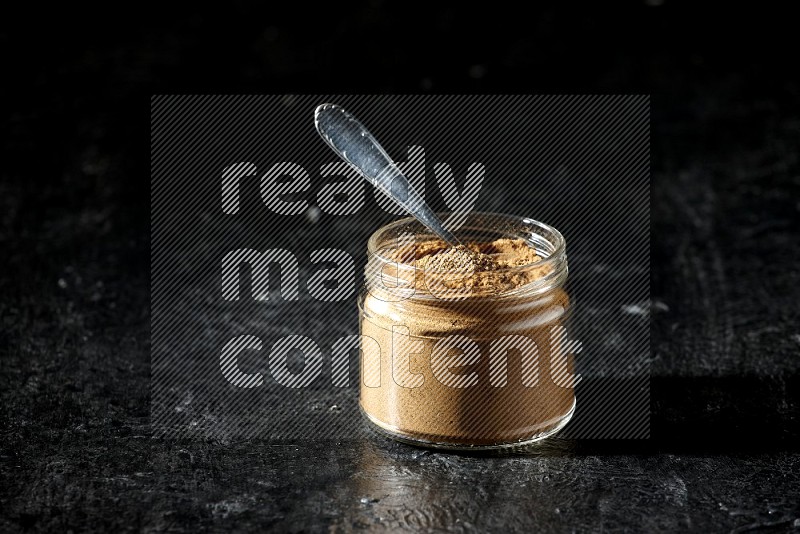 وعاء زجاجي للتوابل وملعقة معدنية ممتلئان ببودرة بهار الفلفل الحلو على خلفية سوداء