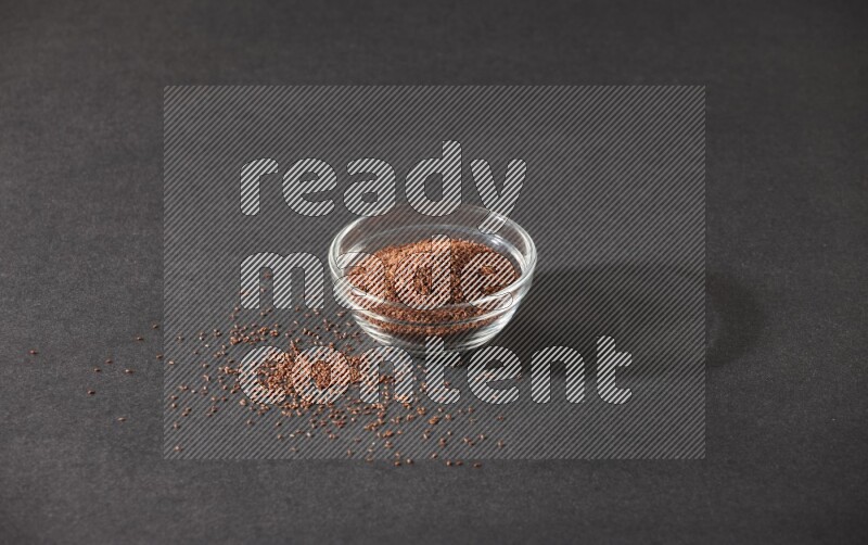A glass bowl full of garden cress seeds surrounded by seeds on a black flooring