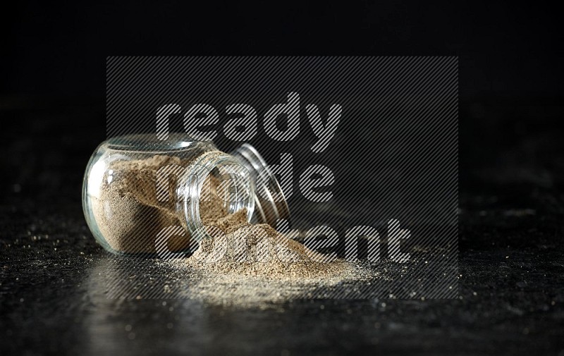 A flipped glass spice jar full of cardamom powder on textured black flooring