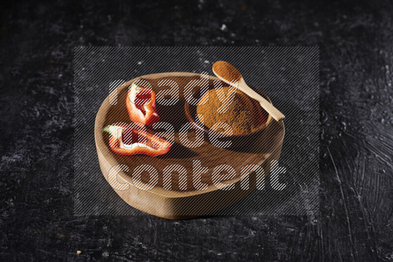 A wooden bowl full of ground paprika powder and sliced red bell pepper beside it, all on a wooden tray on black background