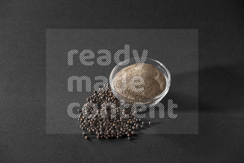 A glass bowl full of powder black pepper powder and black pepper beads on the floor on black flooring