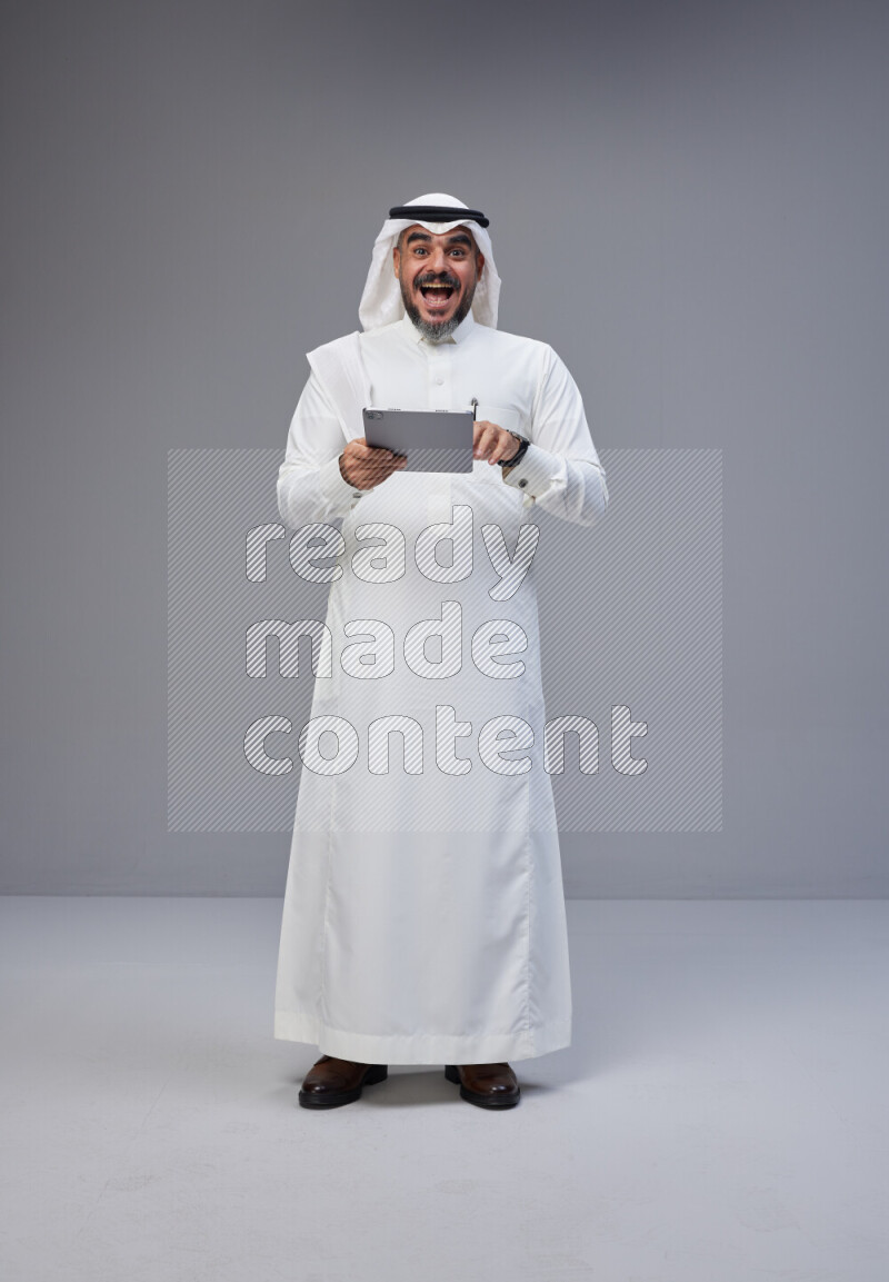 Saudi man Wearing Thob and white Shomag standing working on tablet on Gray background