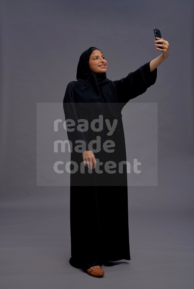 Saudi woman wearing Abaya standing taking selfie on gray background