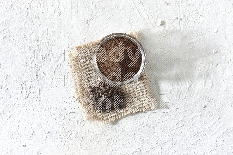 A glass bowl full of cloves powder and cloves grains on a burlap piece on a white flooring
