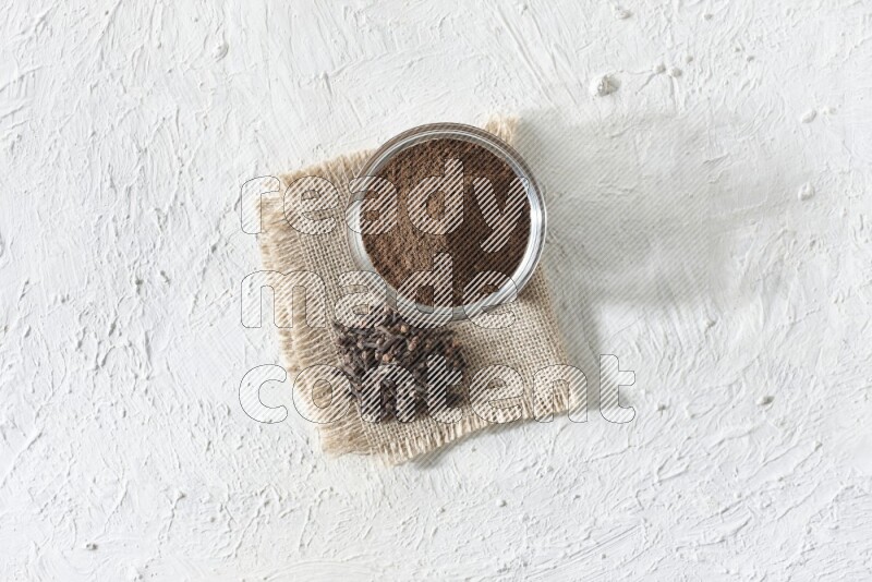 A glass bowl full of cloves powder and cloves grains on a burlap piece on a white flooring